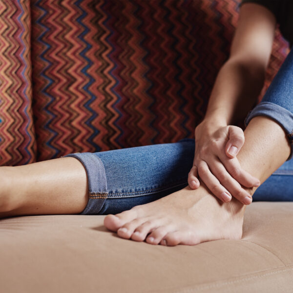 Woman sitting on sofa at home and suffering from leg pain, massaging ankle, body injury